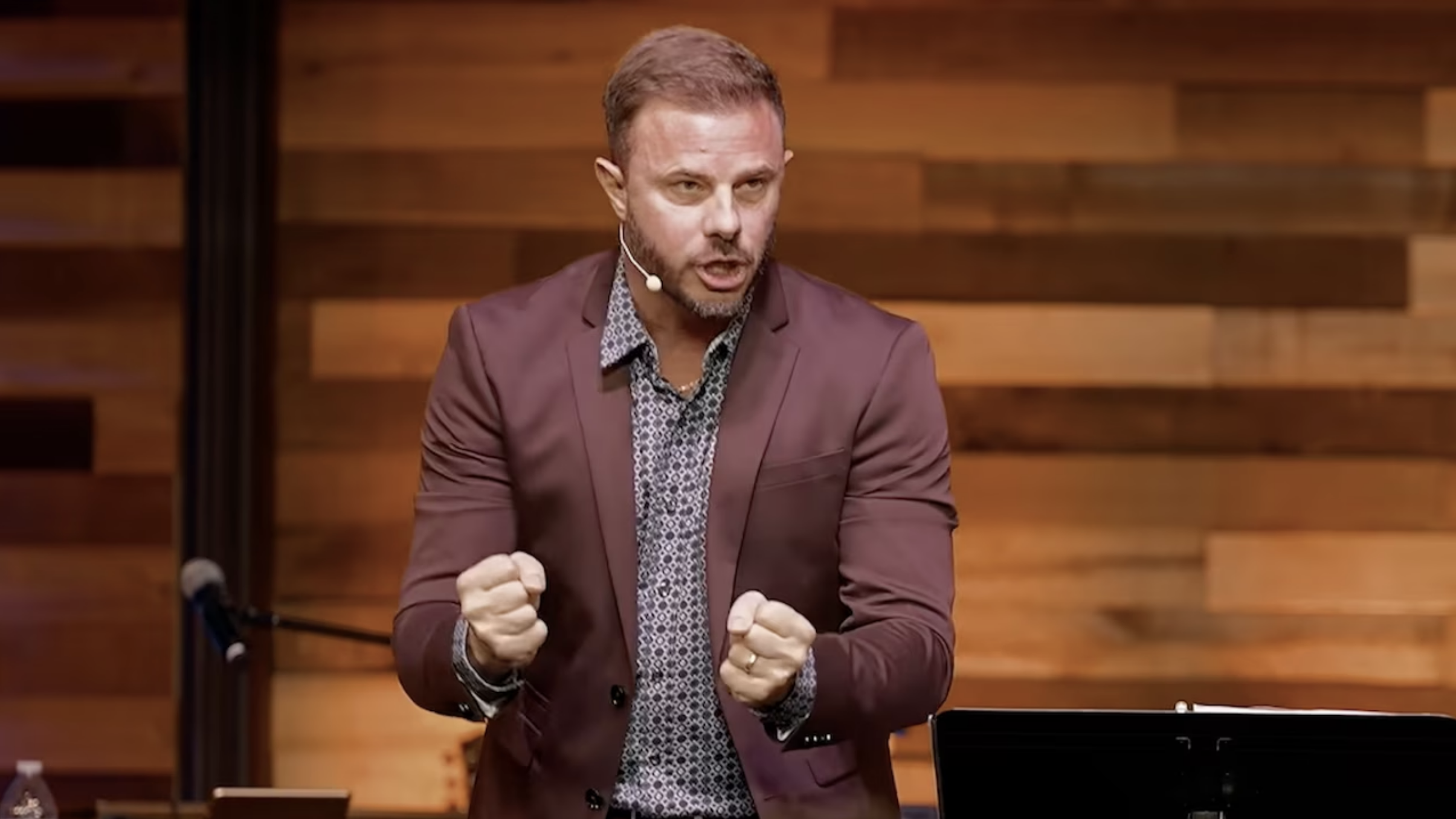 Man in burgundy blazer and patterned shirt speaks passionately on a church stage with a wood-plank wall behind him.