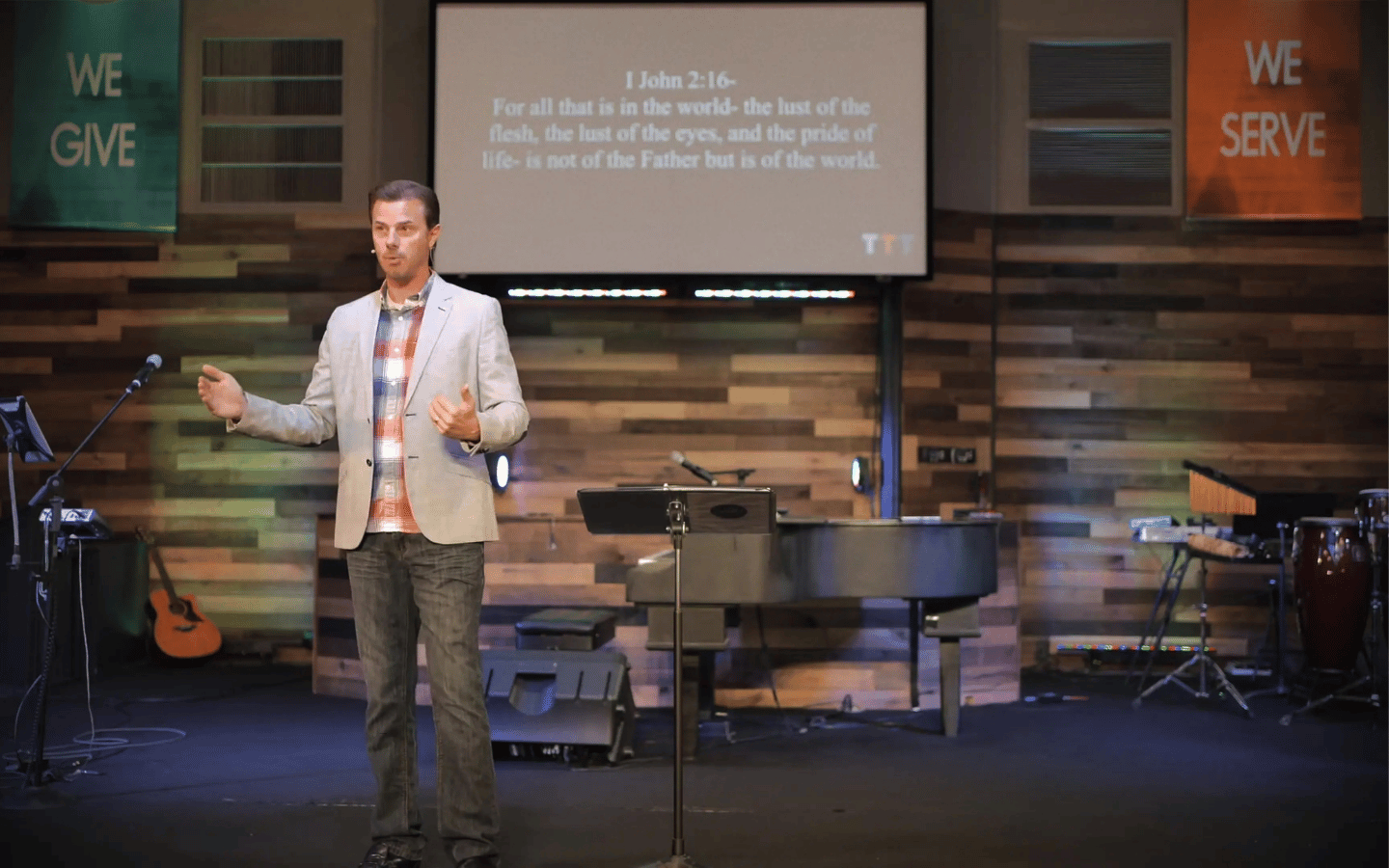 Pastor speaking on church stage with a projection screen showing 1 John 2:16, guitars and drums visible in the background.