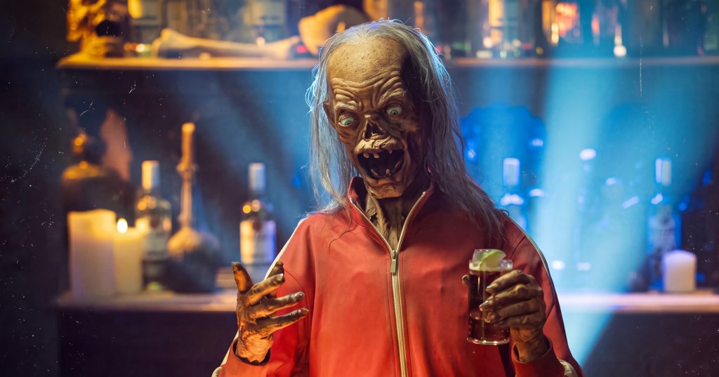 Zombie-like Crypt Keeper puppet in a red track jacket holding a drink behind DJ turntables in a dimly lit bar with skulls and candles in the background.