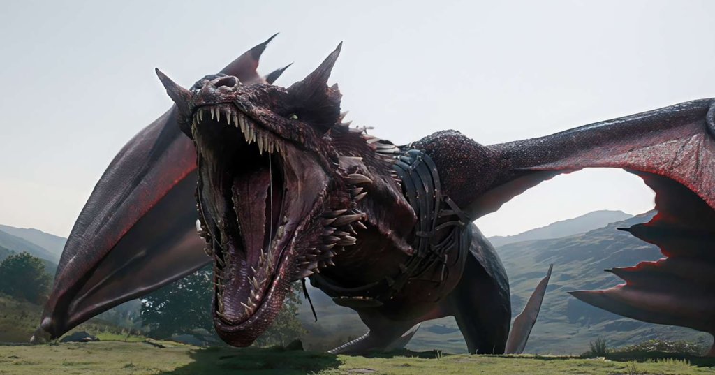 A large dark red dragon with mouth wide open roaring on a green hillside with mountains in the background, wearing a saddle harness.