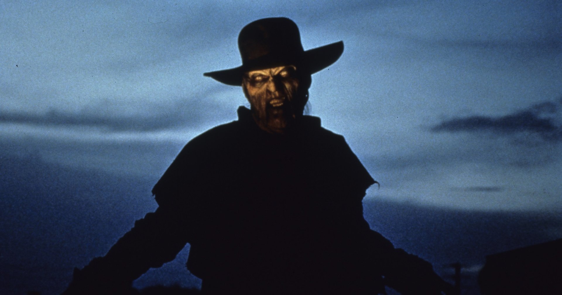 A monstrous figure in a wide-brimmed hat with a decayed, snarling face is silhouetted against a dark blue dusk sky in a horror film still.