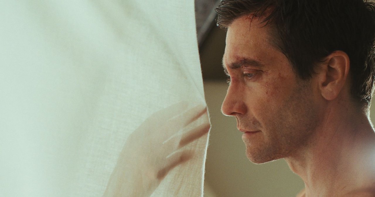 Close-up profile of a shirtless man with dark hair gazing downward beside a sheer white curtain.