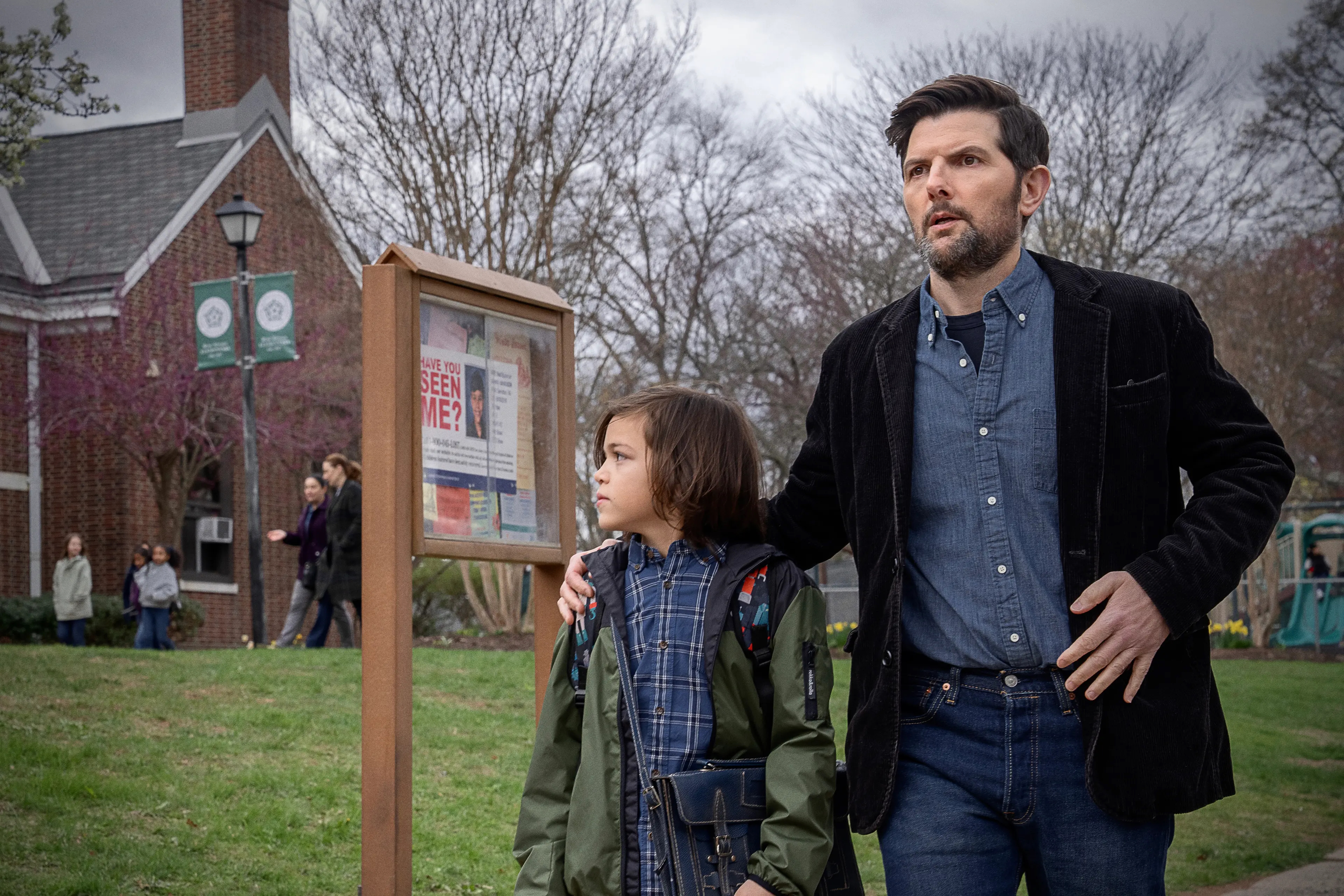 A bearded man in a black jacket and denim shirt stands behind a young boy near a bulletin board with a missing person flyer outdoors.