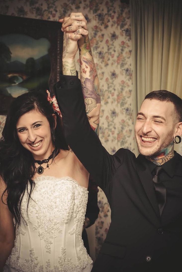 A tattooed bride in a white strapless gown and groom in a black suit raise their clasped hands and laugh joyfully at their wedding reception.