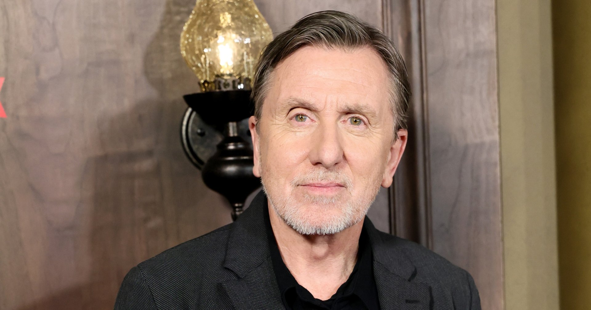 Tim Roth in a dark grey blazer and black shirt, hands in pockets, posing at a premiere event in front of a wooden backdrop with a vintage wall lamp.