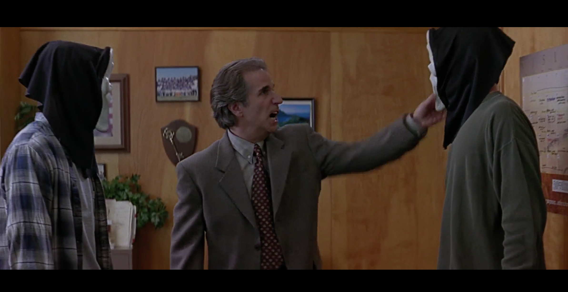 A man in a suit and tie stands between two students with black hoods over their heads in a wood-paneled school office, pointing at one of them.