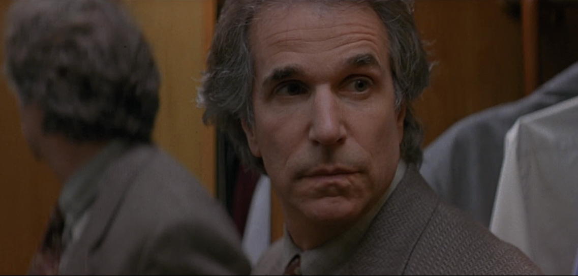 Close-up of a middle-aged man in a suit with a guarded, stern expression, looking over his shoulder in a dimly lit interior.