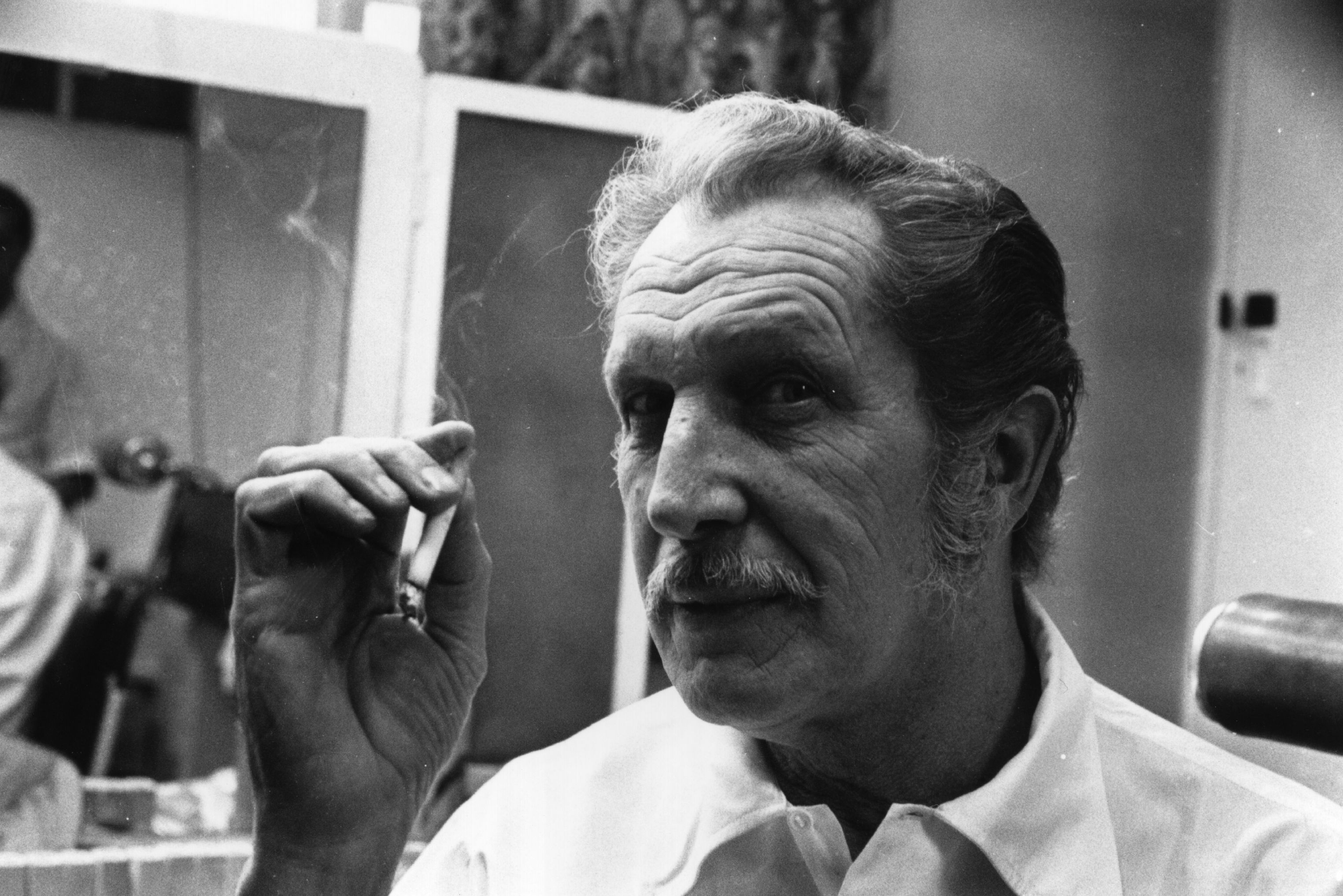 Close-up black-and-white photo of a mustachioed older man smoking a cigarette in a dressing room, reflected in a mirror behind him.