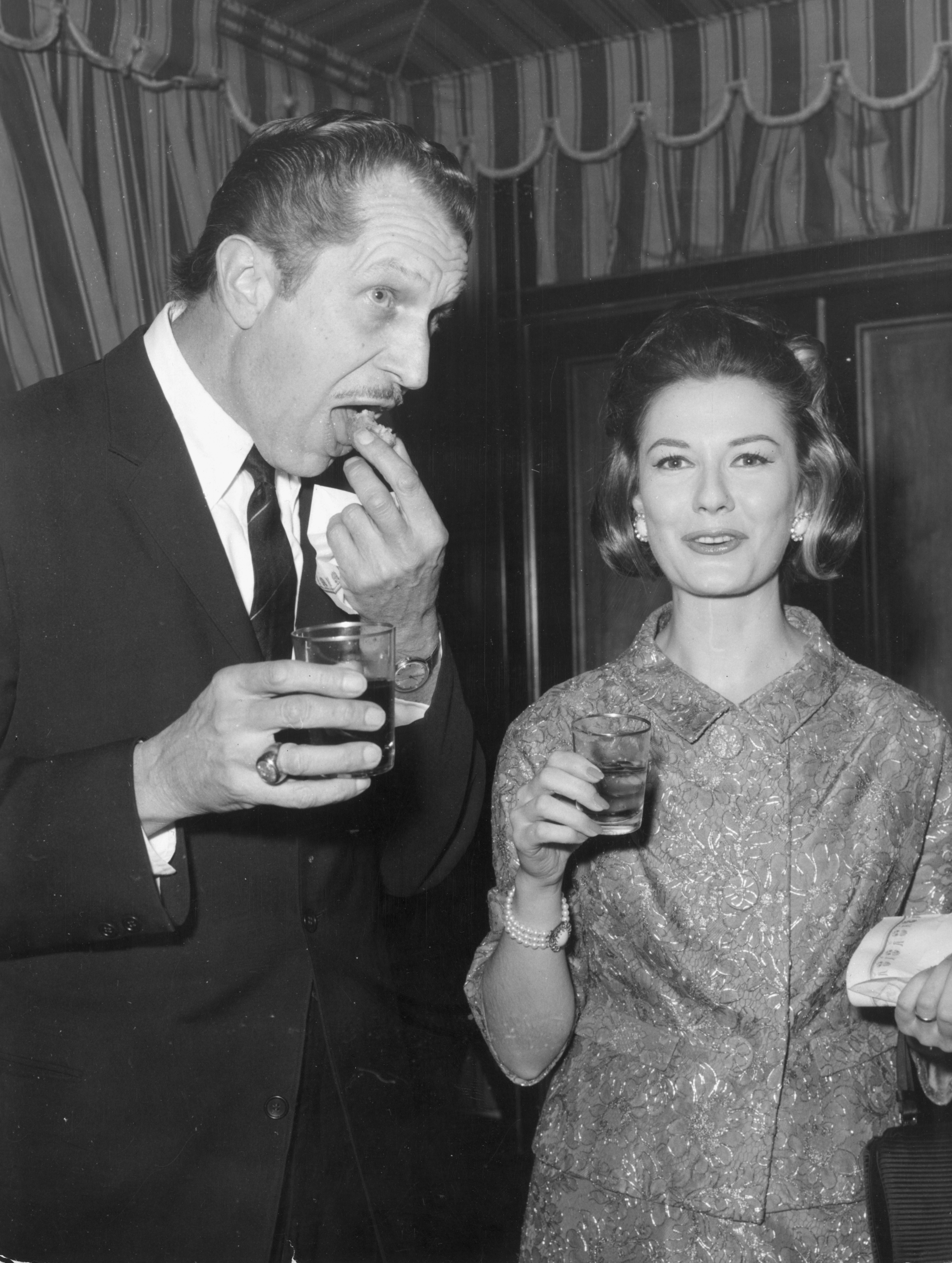 A man eating a snack and holding a drink stands beside a woman in a lace dress also holding a glass, at a cocktail party.