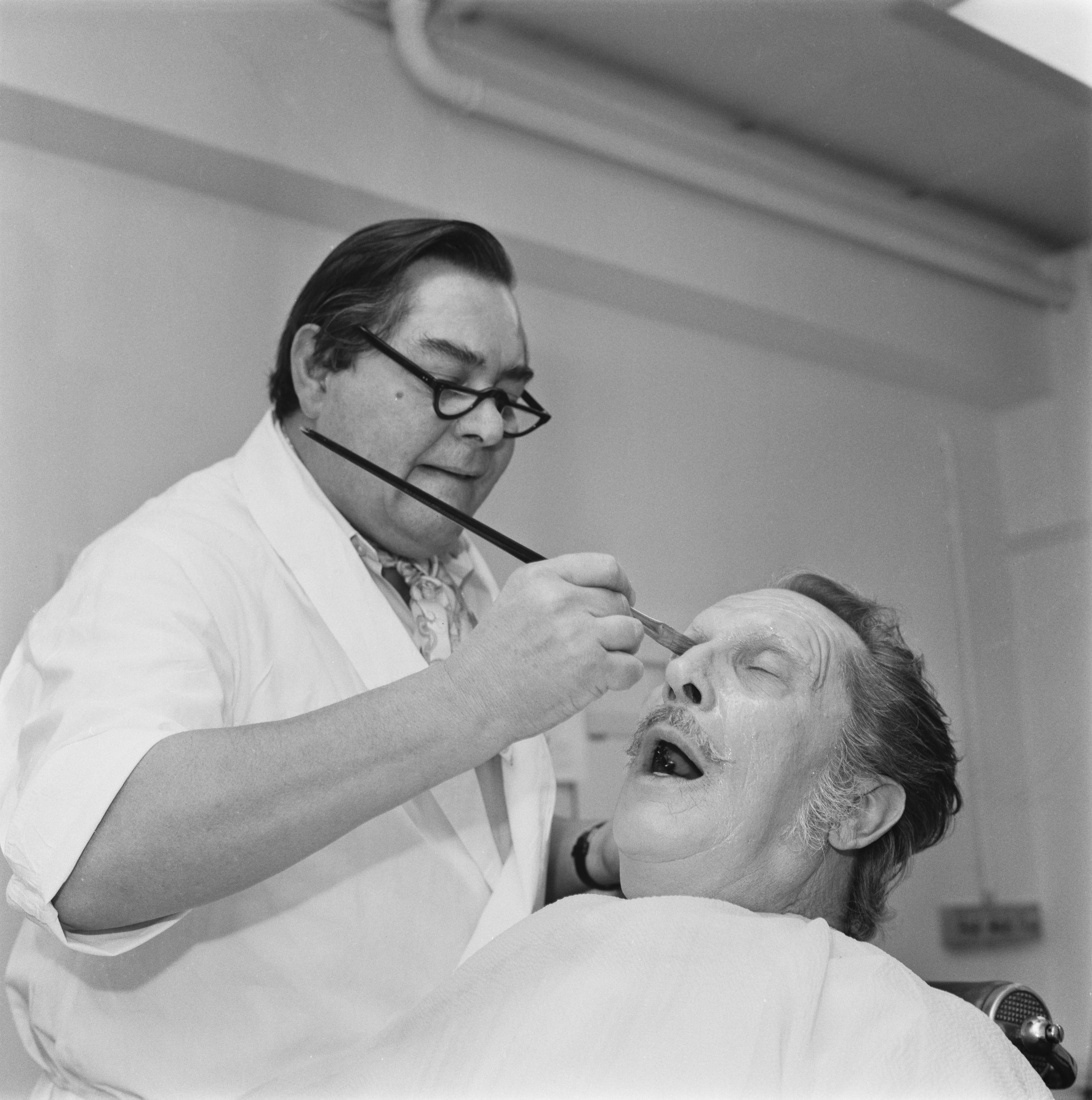 A make-up artist in glasses and a white coat applies make-up with a brush to a reclined man wearing a white cape, whose face is heavily covered in pale prosthetic foundation.