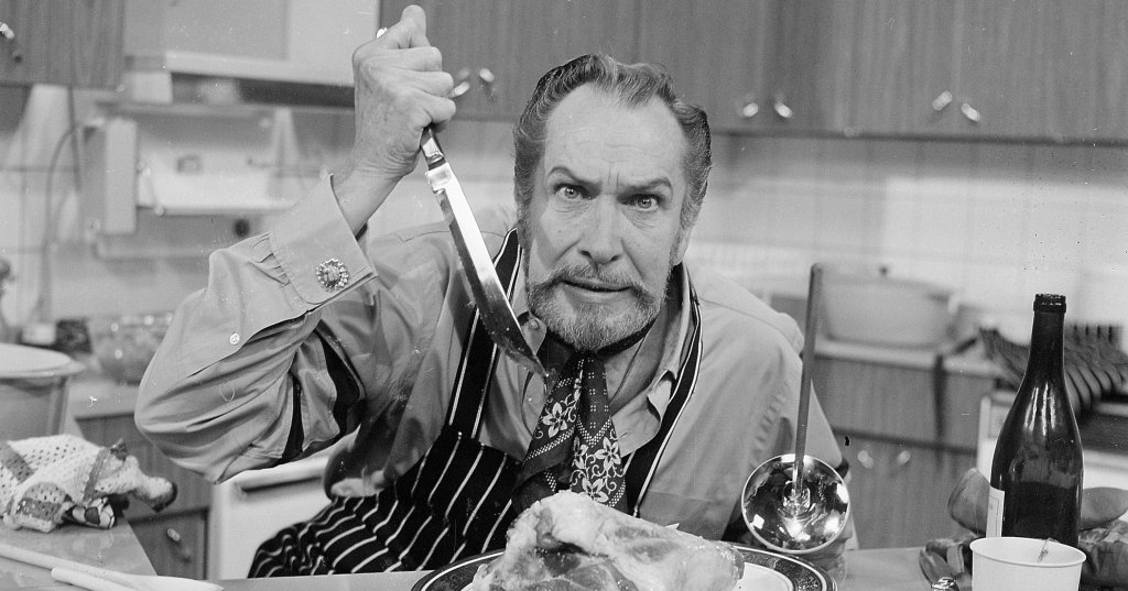 Black-and-white photo of a bearded man in a striped apron and tie holding a carving knife over a large roast on a plate at a kitchen table.