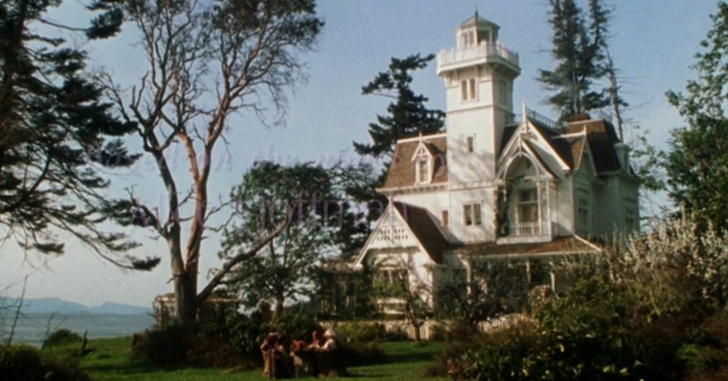 Wide exterior shot of a large white Victorian mansion with a tall tower, surrounded by trees, with mountains visible in the background.