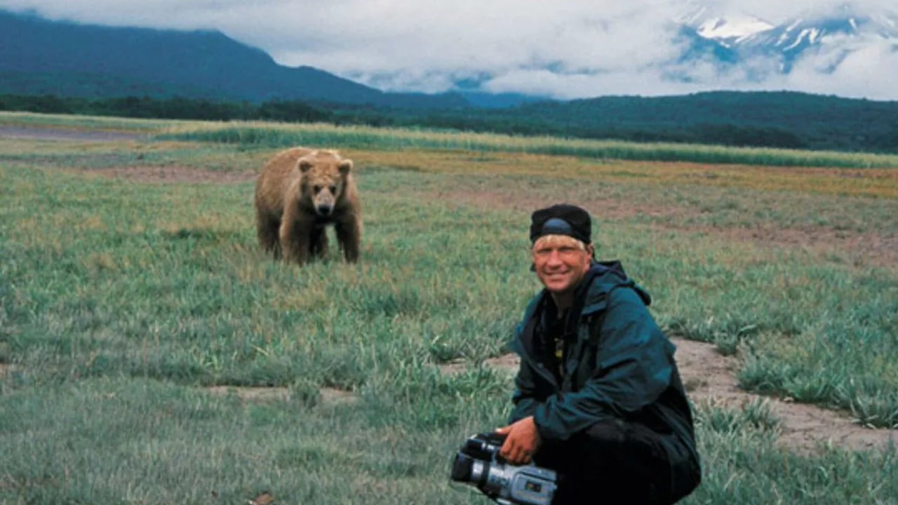 A man crouching with a video camera smiles at the camera while a grizzly bear stands in the field behind him, mountains in the distance.