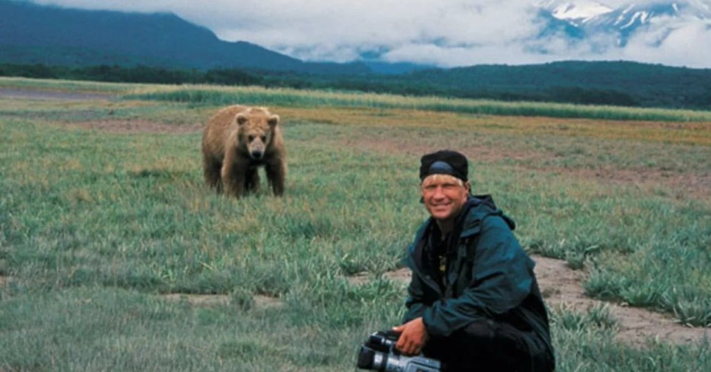 A man crouching with a video camera smiles at the camera while a grizzly bear stands in the grassy field behind him, mountains in the background.