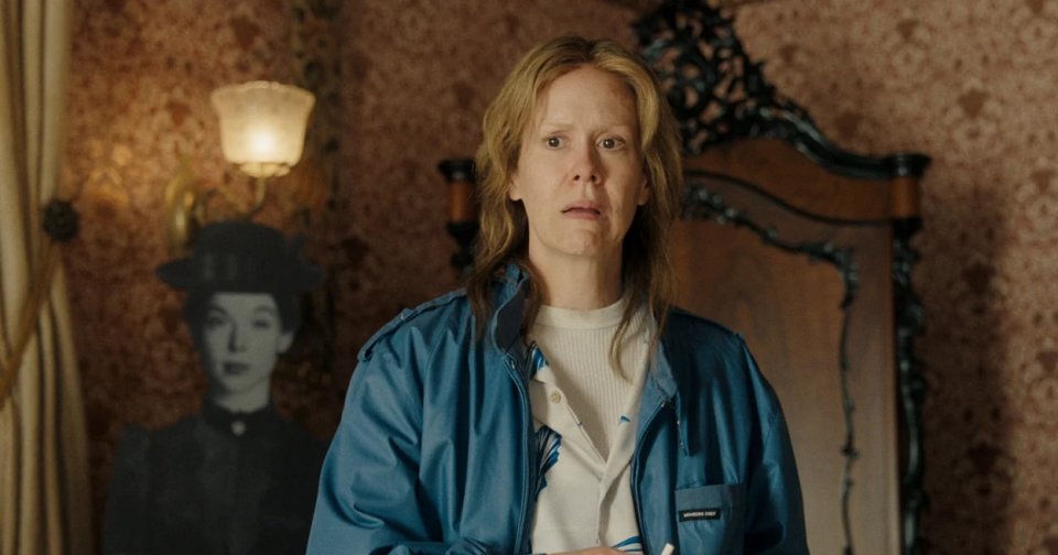 A woman in a blue Members Only jacket holds a cigarette inside a Victorian-wallpapered room, with a shadowy black-and-white figure visible behind her.
