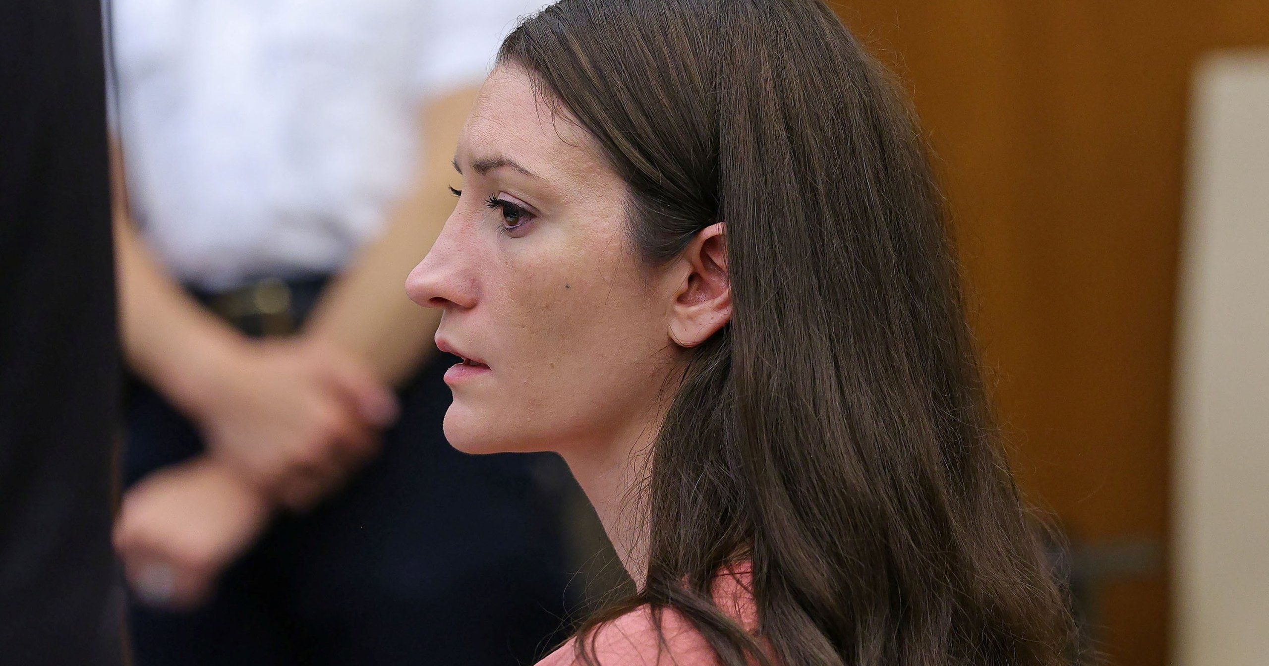 Woman with long brown hair in a pink top seated at a courtroom table, viewed from the side, with a uniformed officer visible in the background.