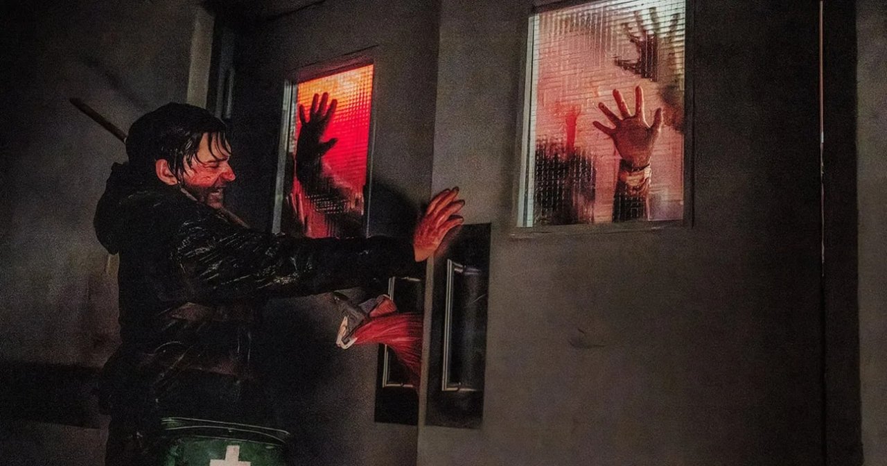 A bloodied man in a black jacket reaches toward a door while zombie hands press through two small red-lit windows in a dark corridor.