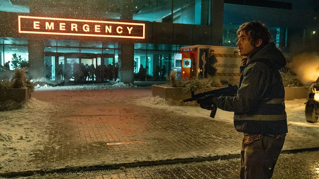 A young man holding a submachine gun stands outside a snow-covered hospital emergency entrance at night, with an ambulance and crowd visible behind him.