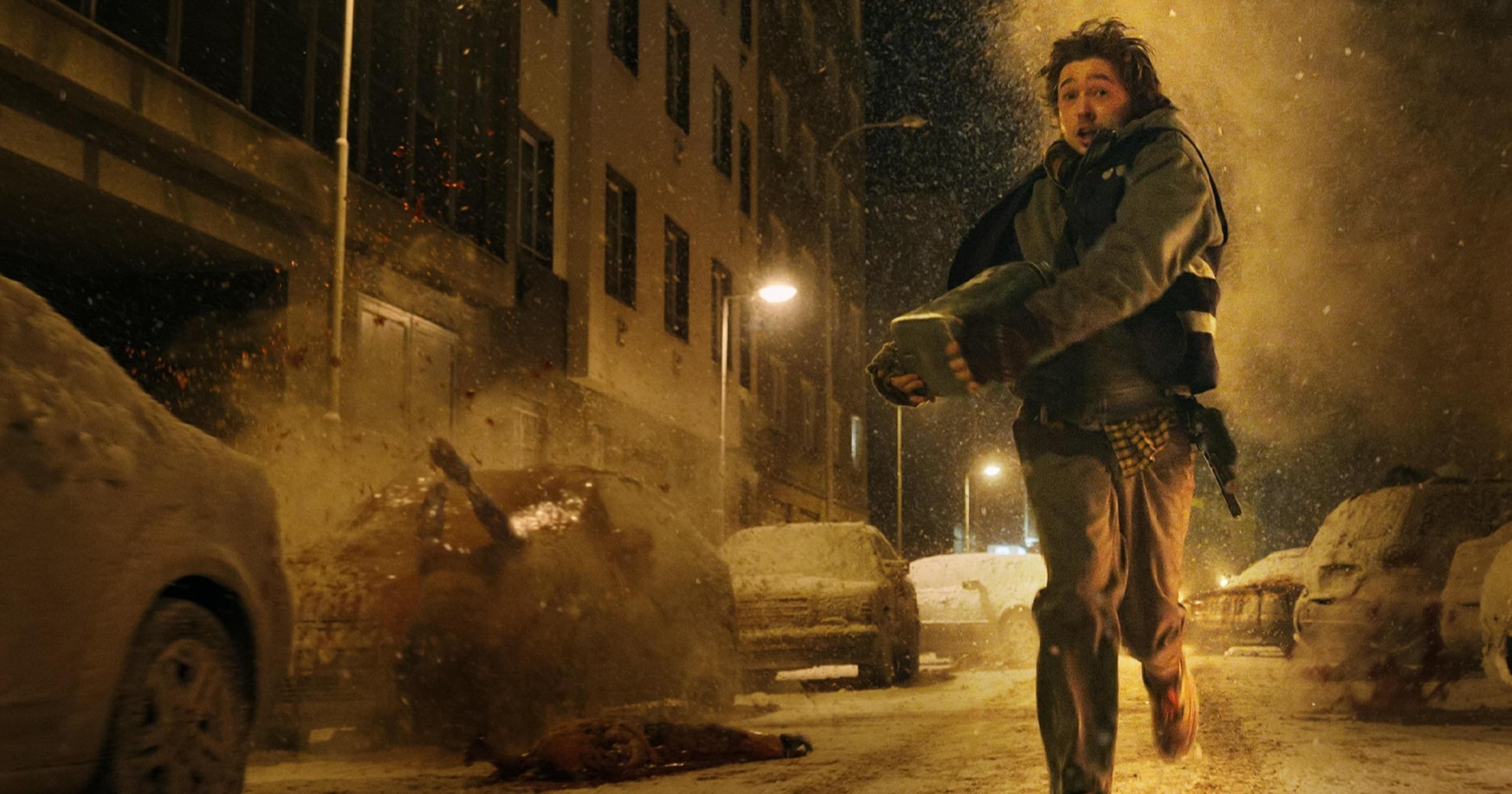A young man runs through a snowy urban street at night, clutching an object, with fire and debris exploding behind him and snow-covered cars lining the road.