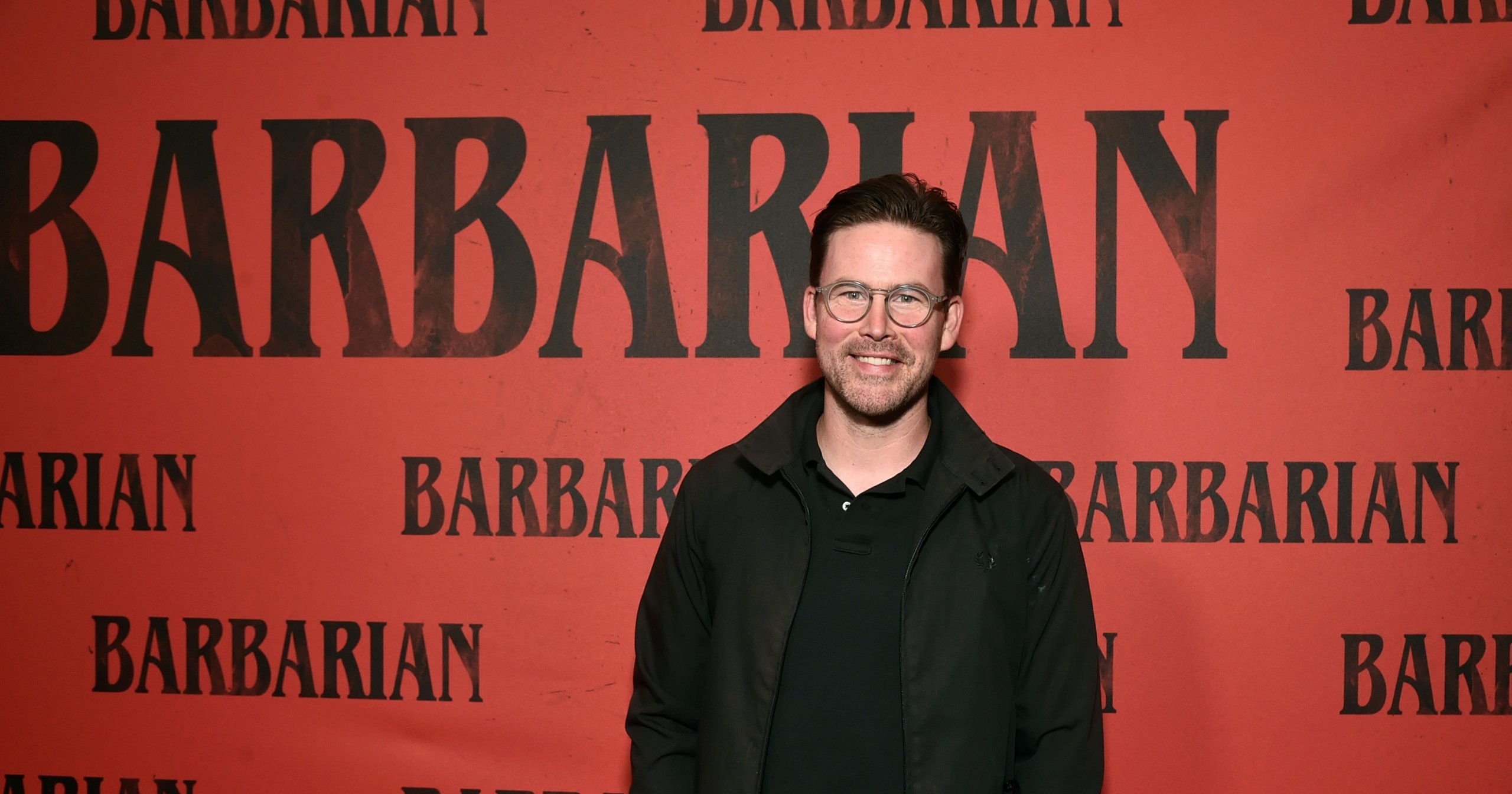 Man in black jacket and glasses smiling in front of a red Barbarian movie promotional backdrop at Comic-Con 2022.