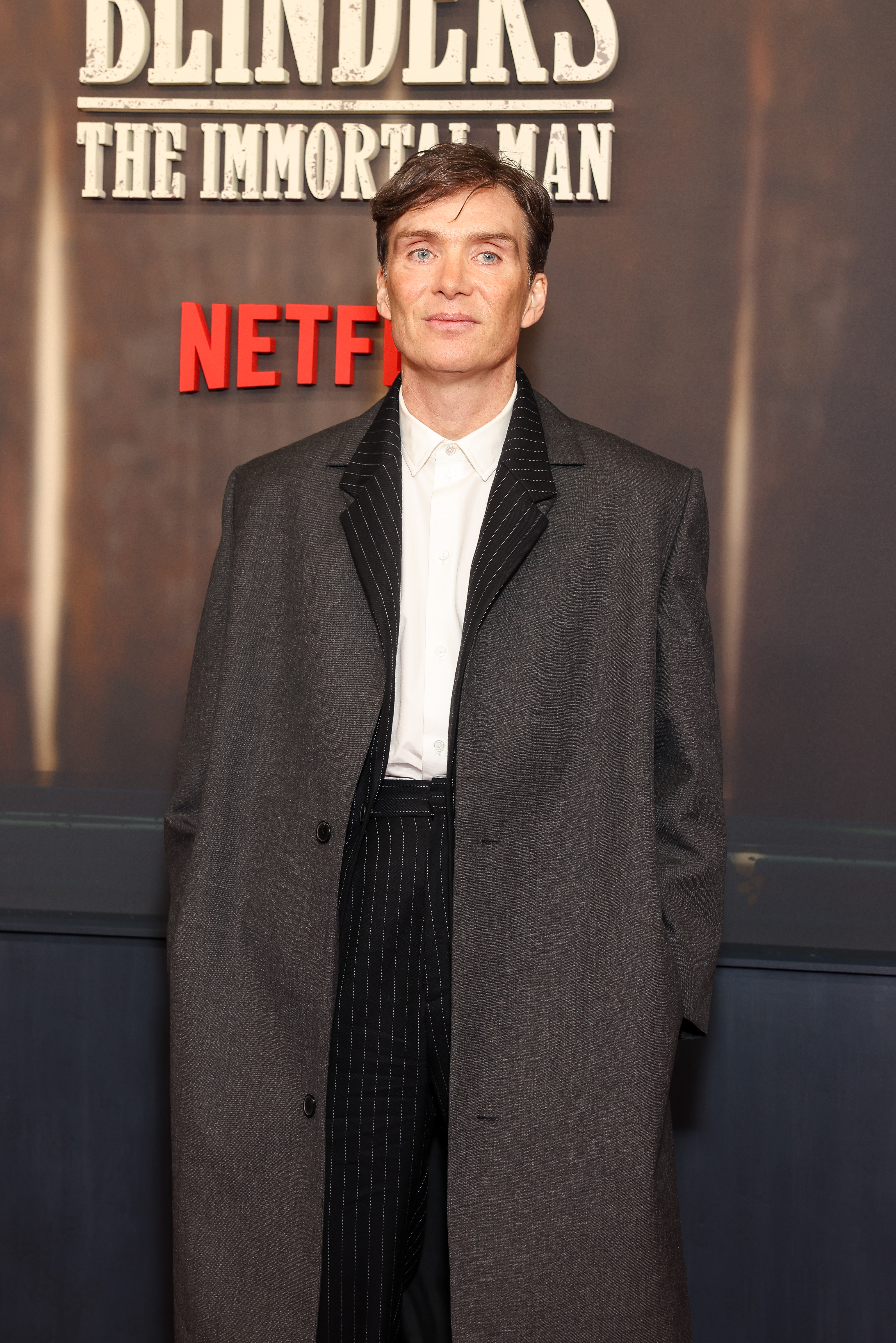 Cillian Murphy in a grey overcoat and pinstripe trousers standing in front of a Peaky Blinders: The Immortal Man Netflix backdrop in Berlin.