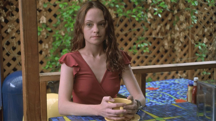 Young woman in a red dress sitting at a colorful mosaic table holding a yellow bowl, looking directly at the camera.