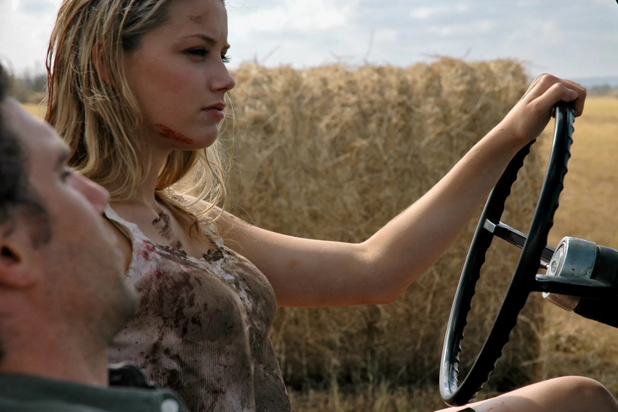 Young blonde woman with blood on her face and torn clothes gripping a tractor steering wheel in a dry field with a hay bale behind her.