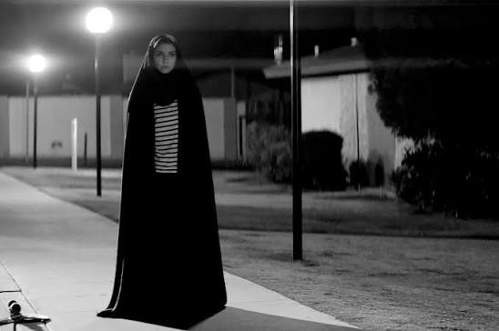 Black-and-white image of a woman in a black chador standing alone on a quiet street at night under a bright streetlamp.