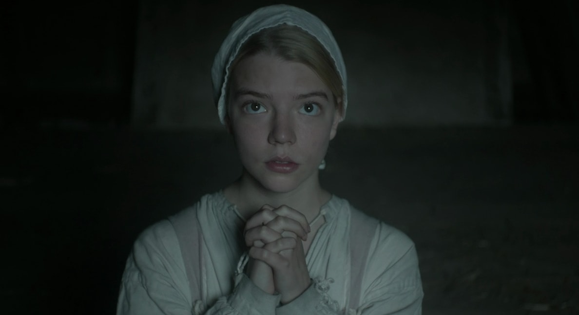 Young blonde girl in a white Puritan dress and bonnet with hands clasped in prayer, looking straight ahead in a dark room.