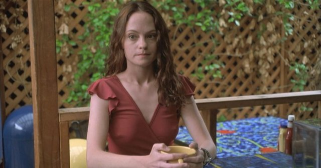 Young woman with long red hair in a red dress sitting at a mosaic outdoor table, holding a yellow bowl and staring forward.