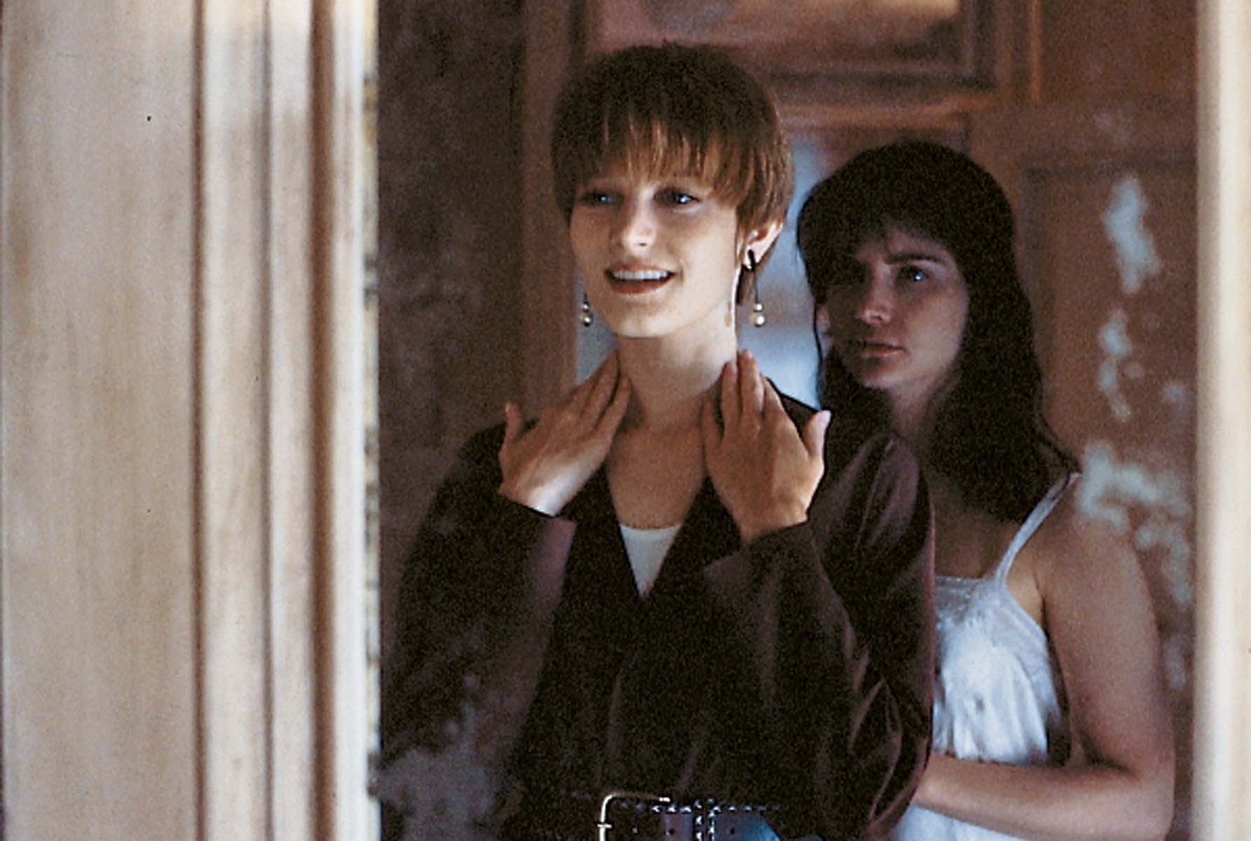 Click to open the image gallery Two women seen through a doorway: a short-haired brunette smiling while a dark-haired woman behind her places hands on her neck near a mirror.
