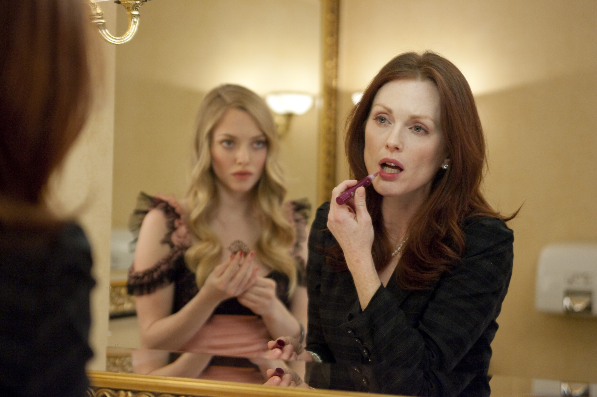 Click to open the image gallery A red-haired woman applies lipstick at a gold-framed bathroom mirror while a young blonde woman in a black dress is visible in the reflection behind her.