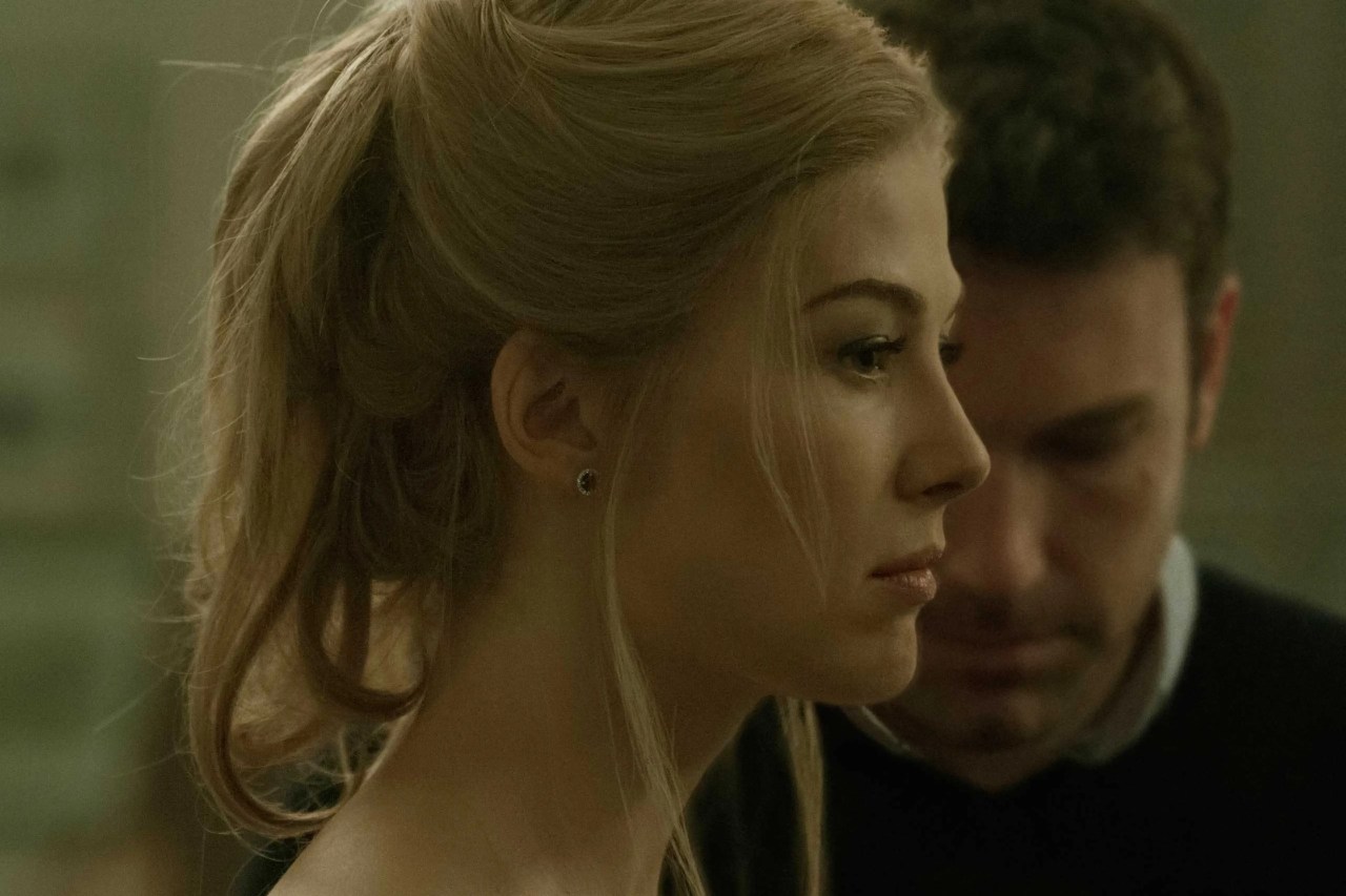 Close-up profile of a blonde woman with her hair in an updo, a dark-haired man blurred in the background behind her, lit in cold green tones.