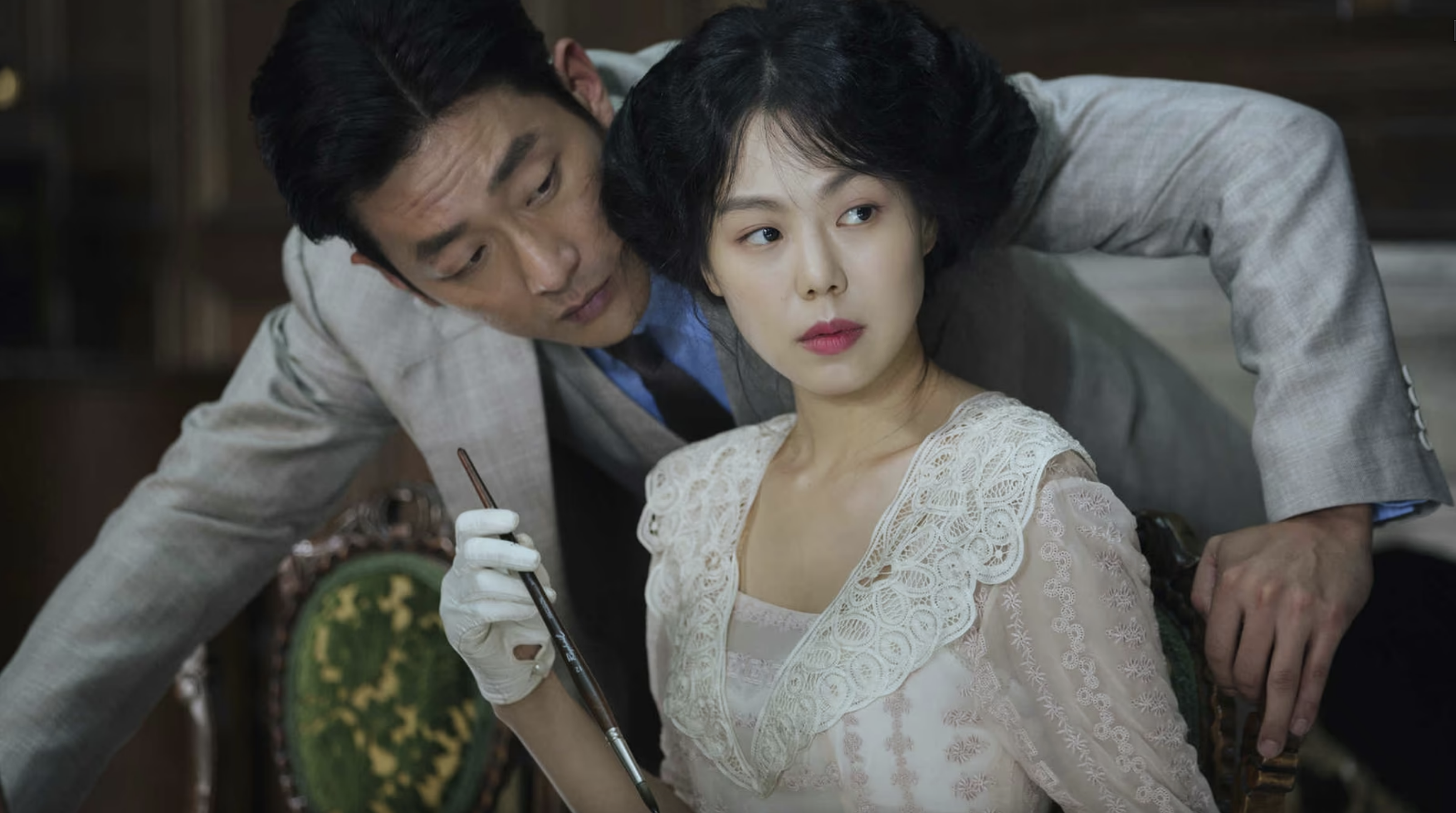 Click to open the image gallery A woman in a white lace dress sits in an ornate chair while a man in a gray suit and white gloves leans over her shoulder, holding a brush.