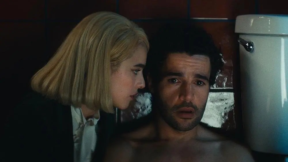 Click to open the image gallery Blonde woman leaning toward a shirtless man sitting against a bathtub in a dimly lit bathroom.