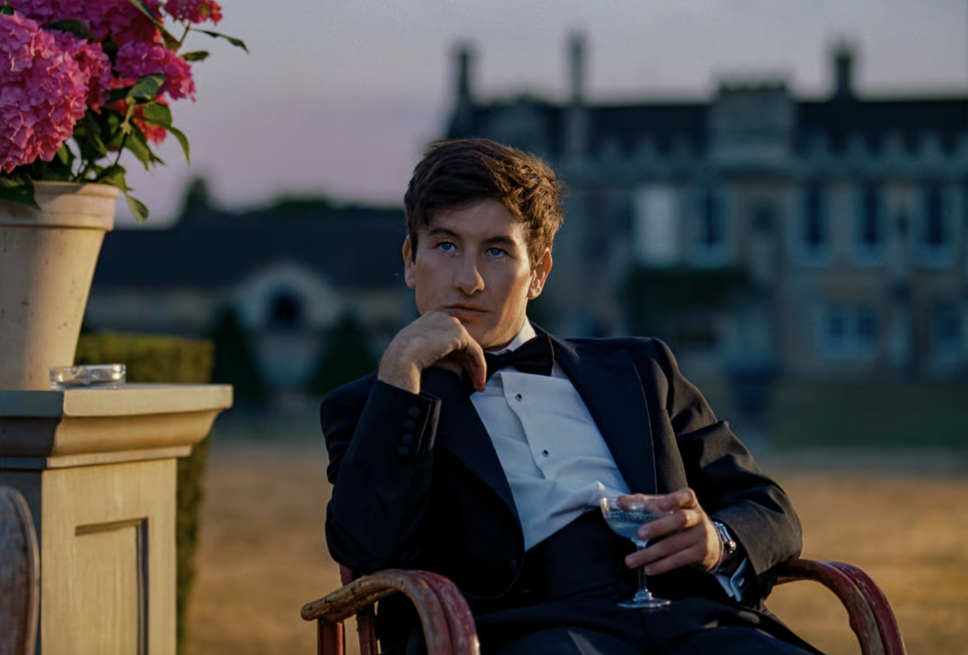 Click to open the image gallery Young man in a tuxedo sitting outdoors in a chair, holding a drink, with a large manor house blurred in the background.