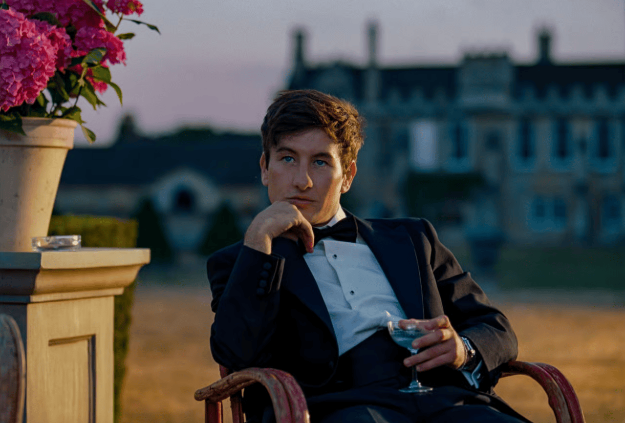 Young man in a tuxedo sitting outdoors in a chair, holding a drink, with a large manor house blurred in the background.