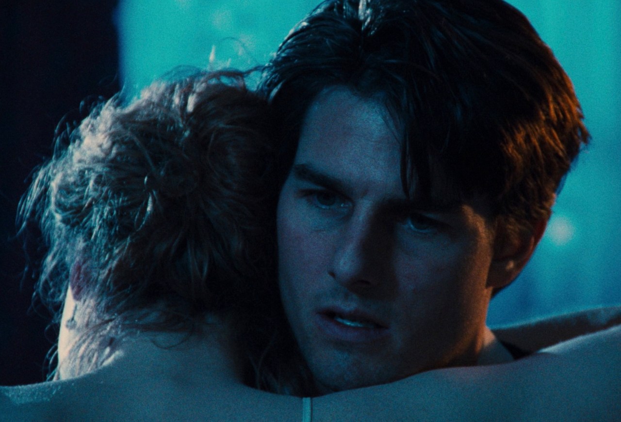A dark-haired man with an intense expression rests his head against someone's shoulder in a dimly lit, blue-toned scene.