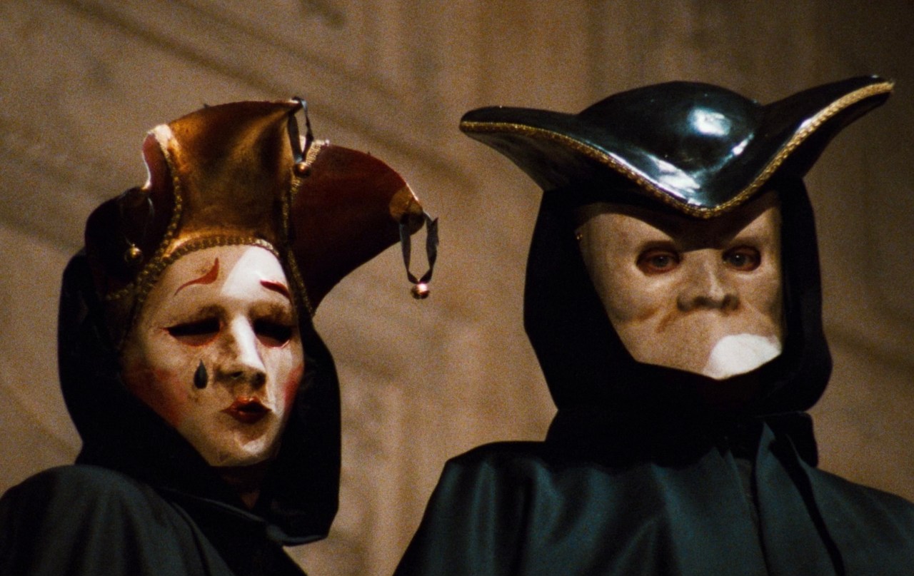 Two figures in black robes and white Venetian masks face forward — one wearing a jester-style hat with a teardrop painted below the eye, the other a black tricorn hat.