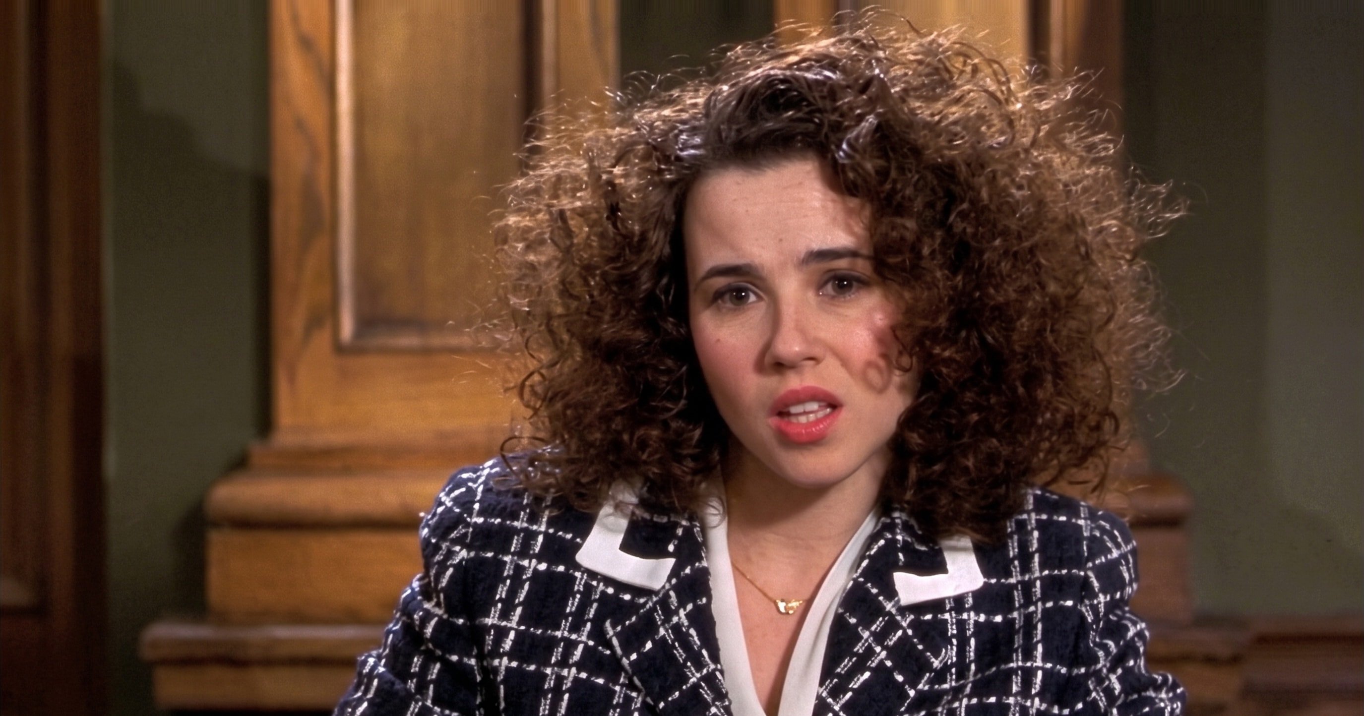 A young woman with voluminous curly hair and a mark on her cheek, wearing a dark plaid blazer, in a scene from Legally Blonde (2001).