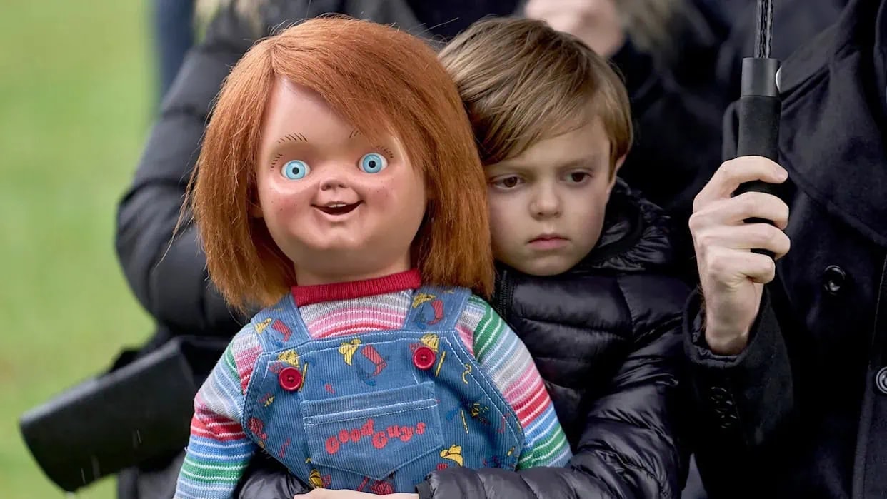 A young brown-haired boy in a dark jacket holds the Chucky Good Guy doll outdoors against a green background.