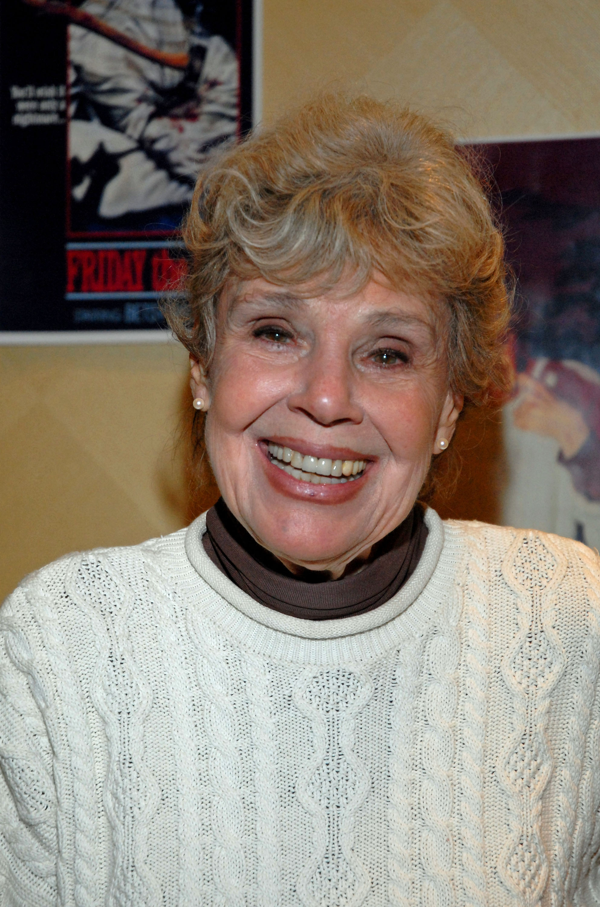 Older woman with short blonde hair smiling in a white cable-knit sweater, a Friday the 13th movie poster visible behind her.