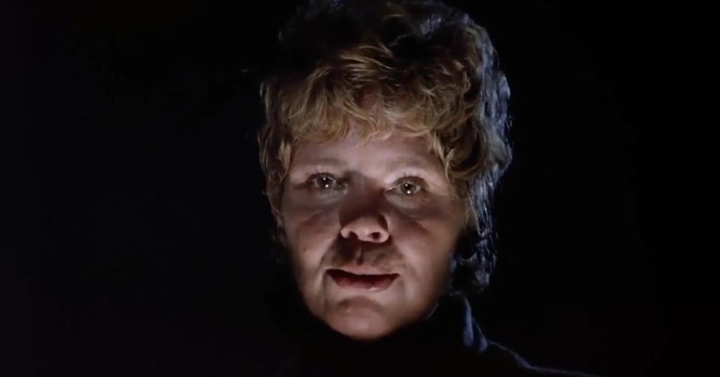 Older woman with short blonde-gray hair wearing a white cable-knit sweater, smiling in front of a Friday the 13th poster.