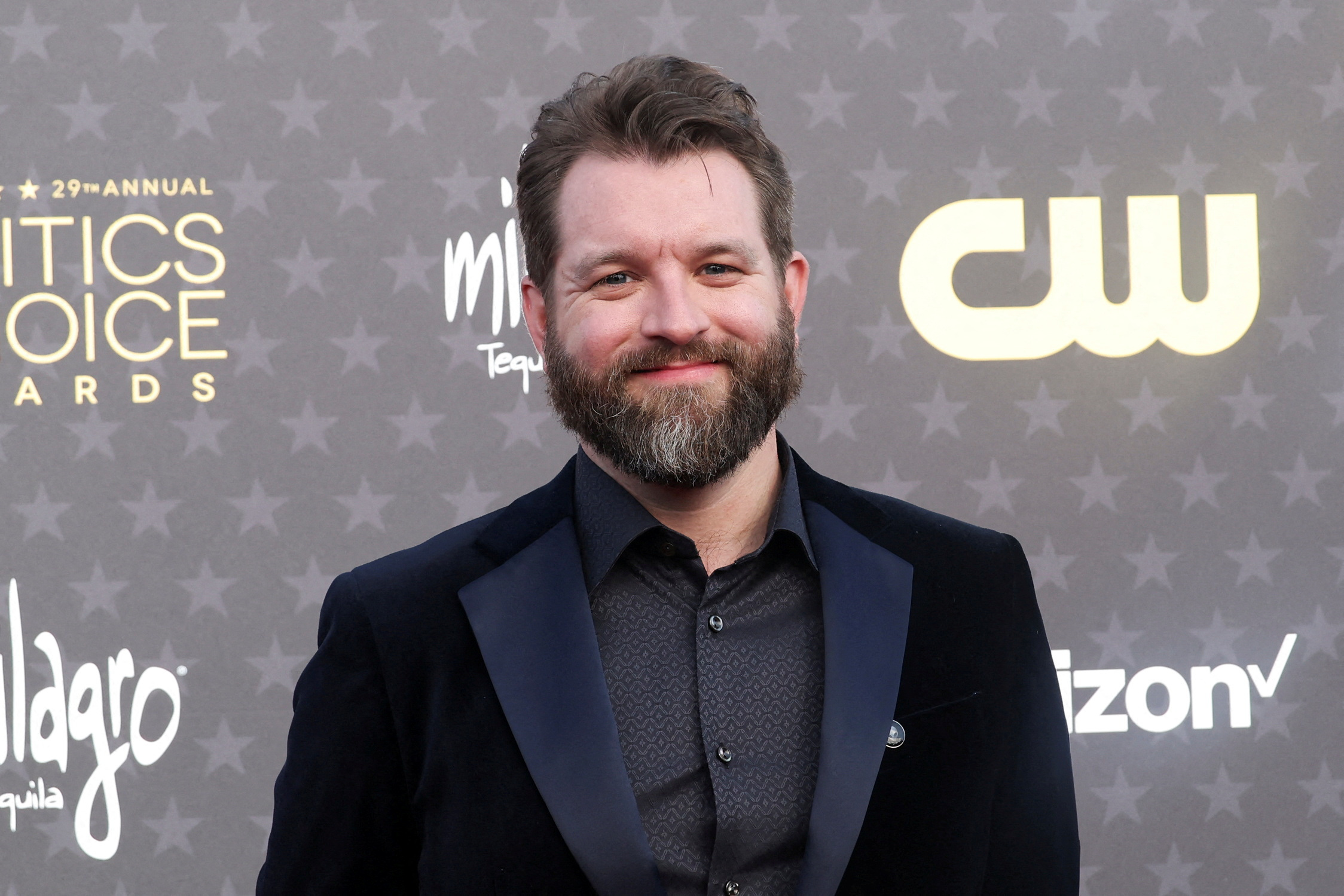 Man with brown hair and beard in a dark blazer smiling at the 29th Annual Critics Choice Awards red carpet.
