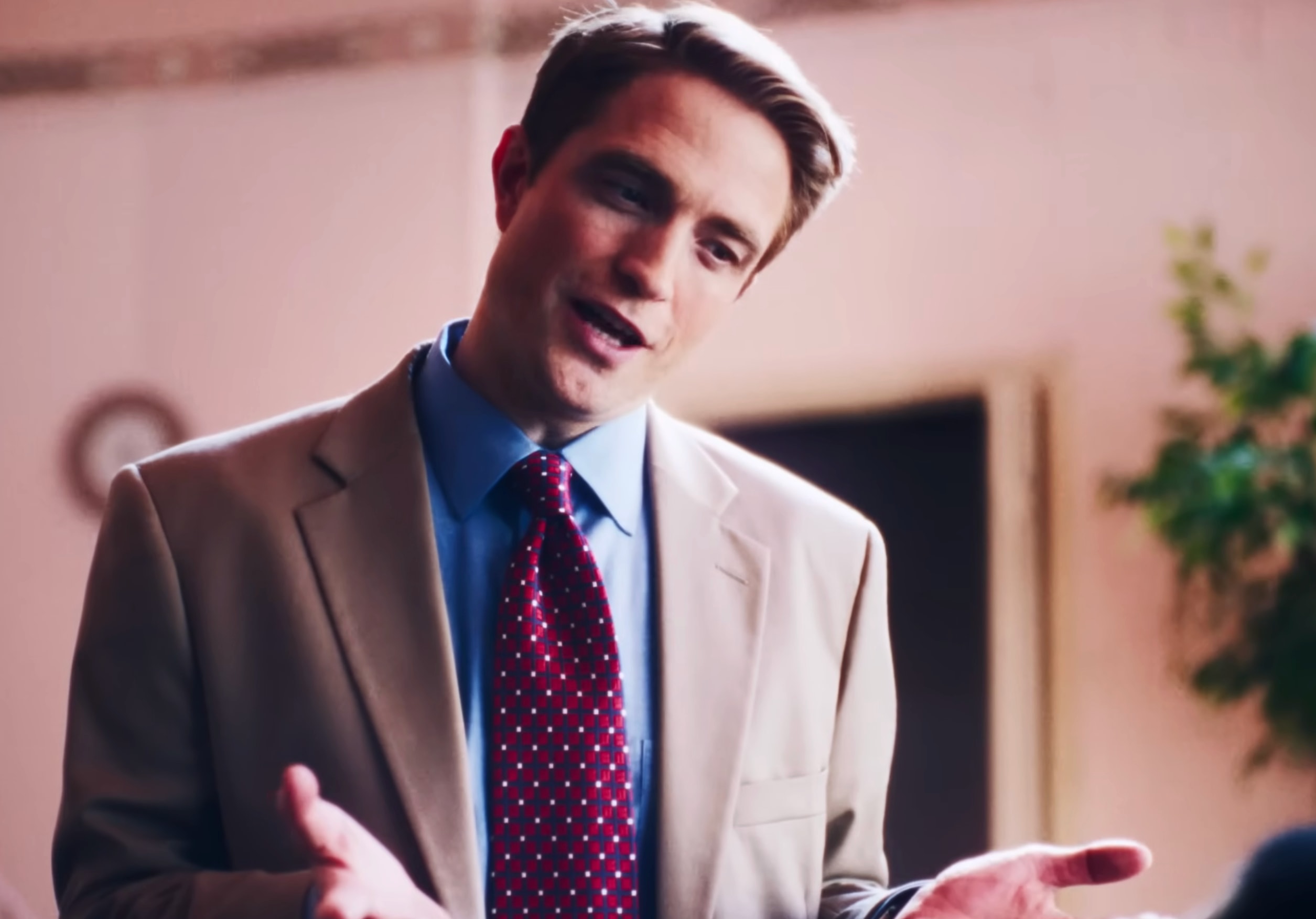 Man in tan blazer and red dotted tie gesturing with both hands while speaking indoors.