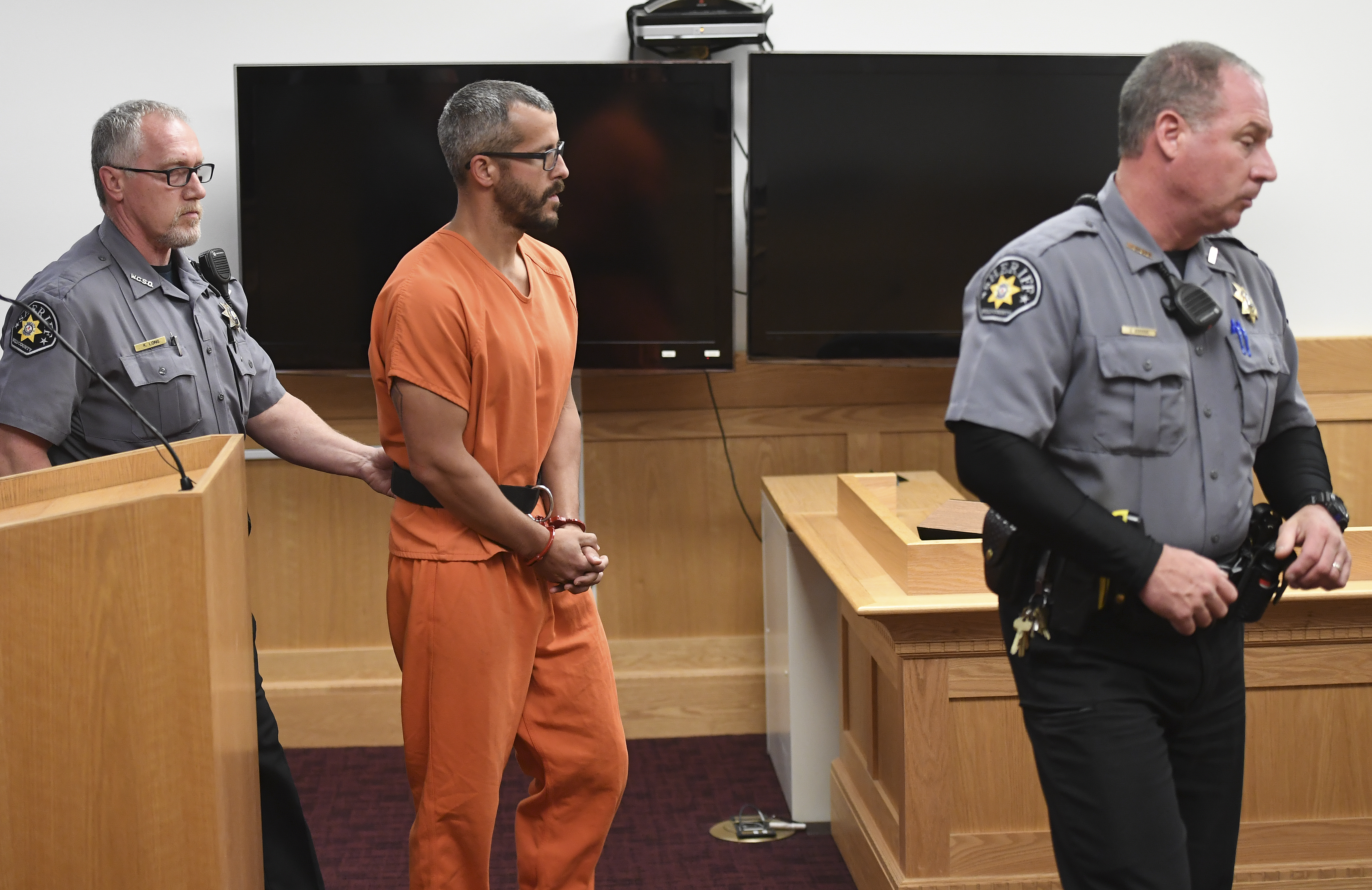Christopher Watts in orange jumpsuit and handcuffs stands between two sheriff's deputies in a courtroom.