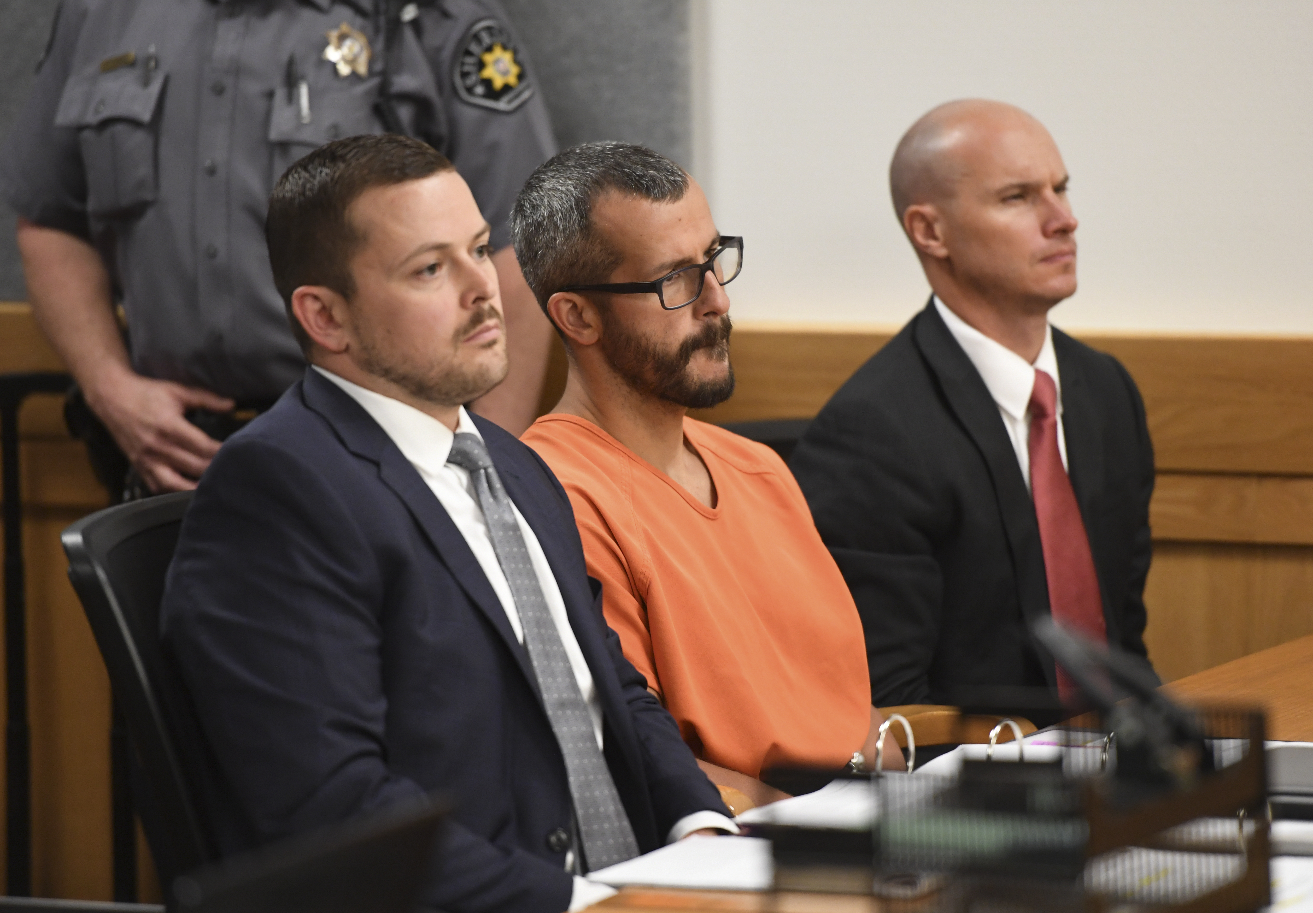 Christopher Watts in an orange jumpsuit sits at a courtroom table between two suited attorneys, a sheriff's deputy visible behind them.