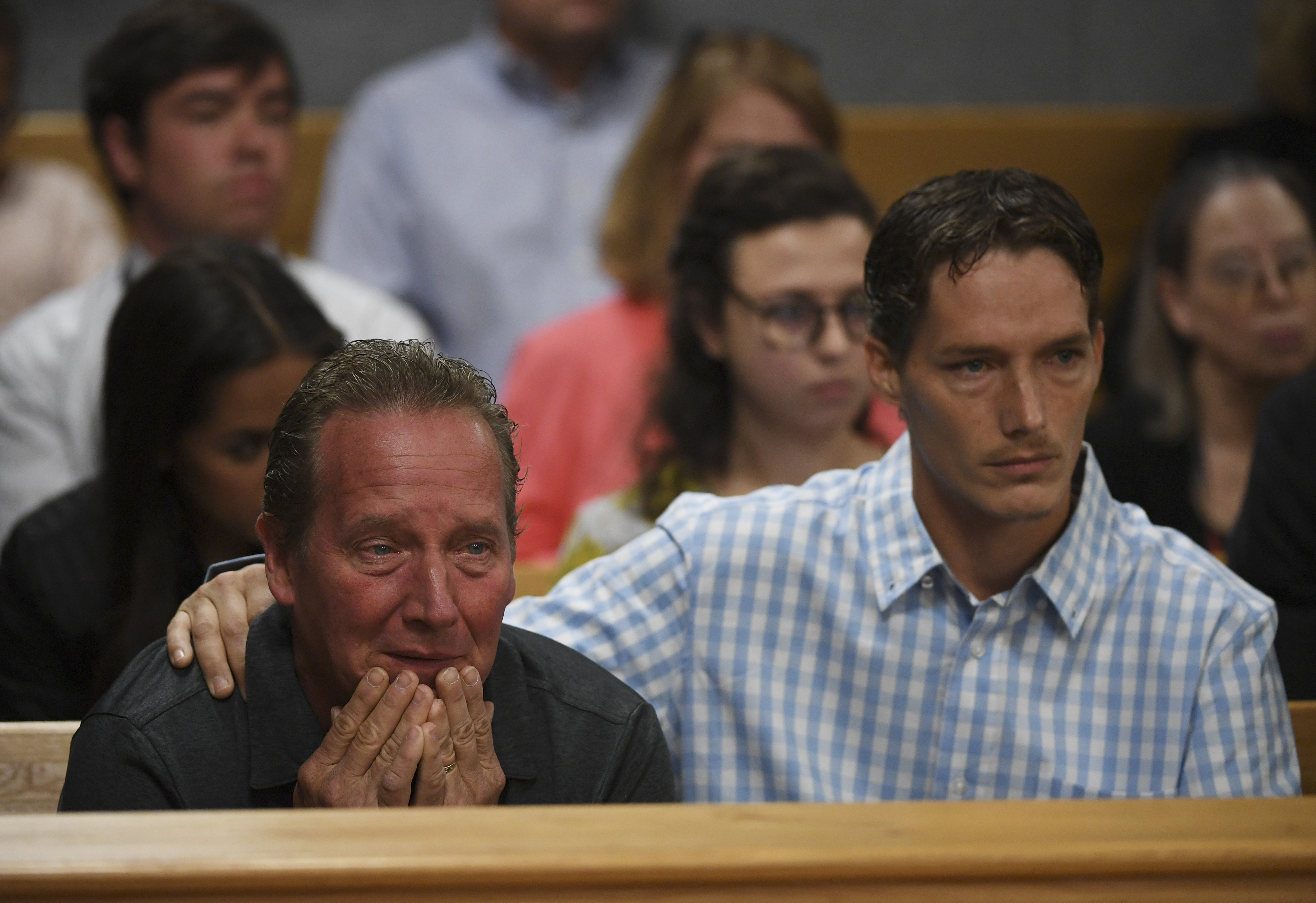 An older man covers his face with his hands in grief while a younger man places a comforting hand on his shoulder in a courthouse gallery.
