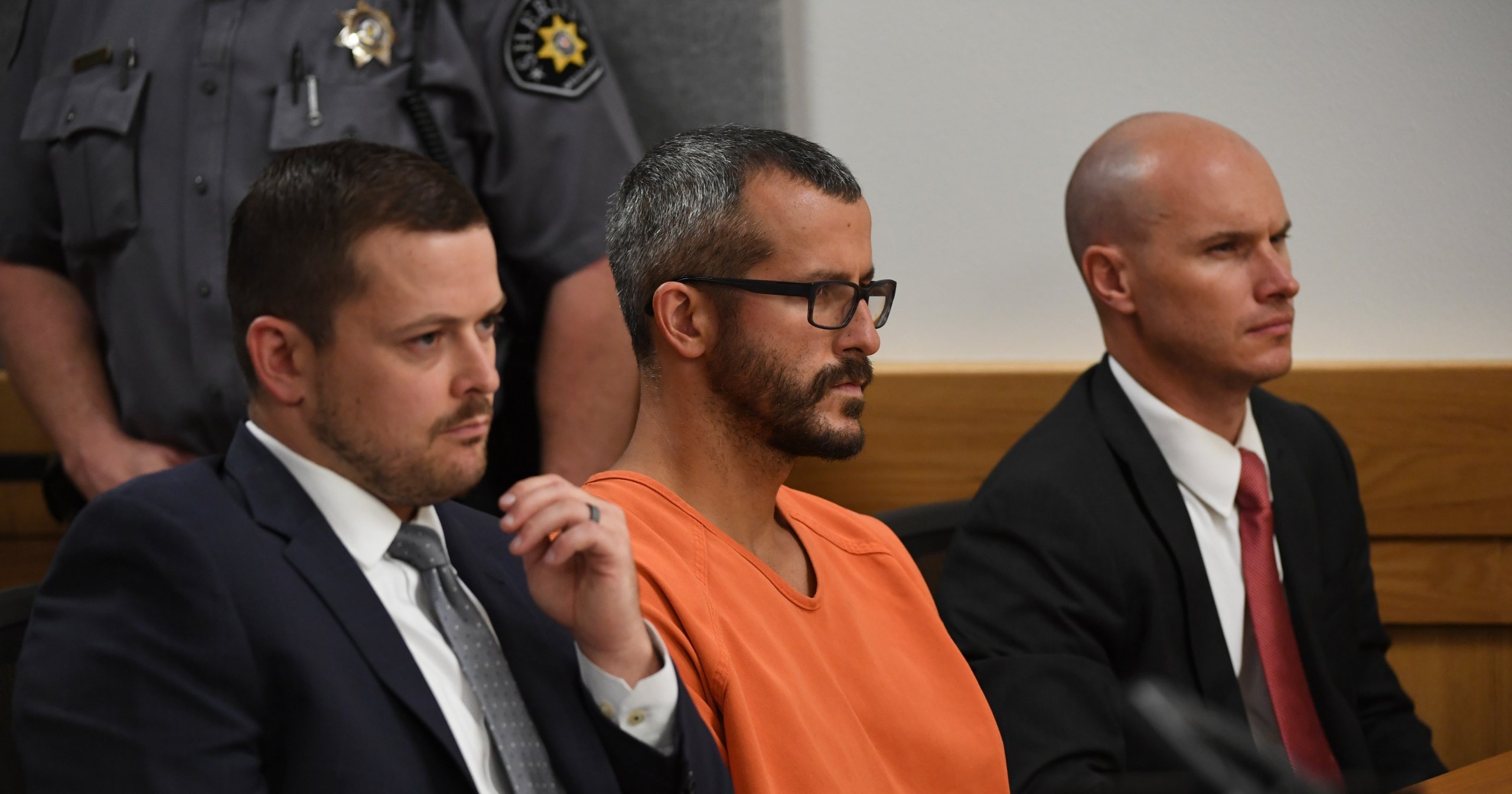 Man in orange jail jumpsuit and glasses seated between two suited attorneys at a courtroom table, with sheriff's deputies standing behind them.