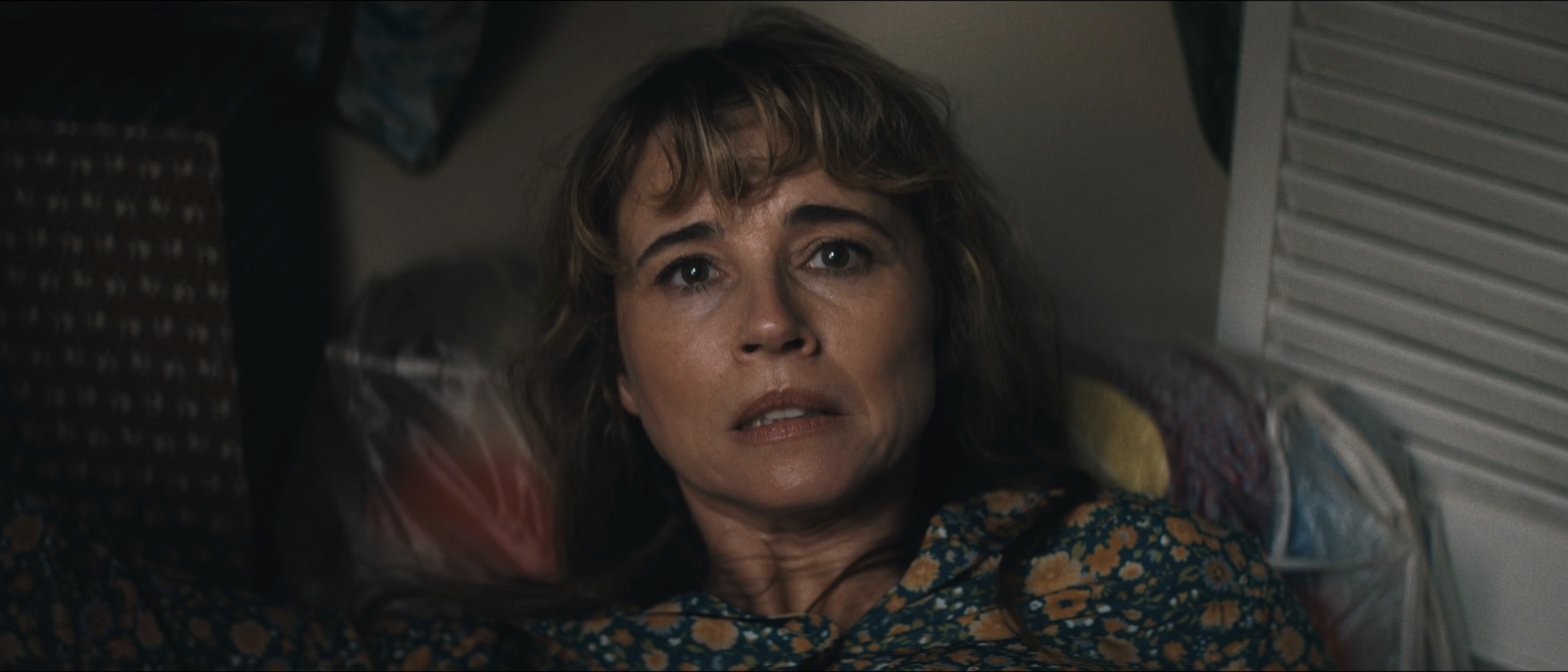 A frightened woman with brown hair in a floral top lying on the floor, looking up anxiously, with louvered closet doors visible beside her.